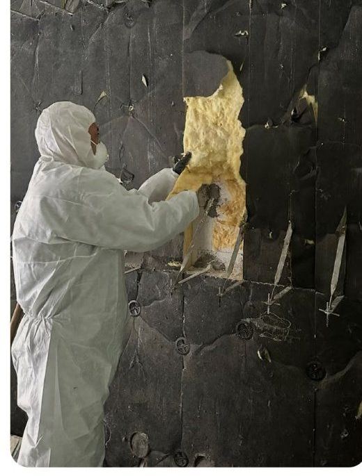 Mitarbeiter im weißen Vollschutzanzug entfernt gelbe Mineralwolle aus einer dunklen Wandkonstruktion