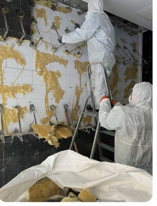 Zwei Mitarbeiter in weißen Schutzanzügen entfernen Dämmmaterial von einer Wand, einer steht auf einer Leiter